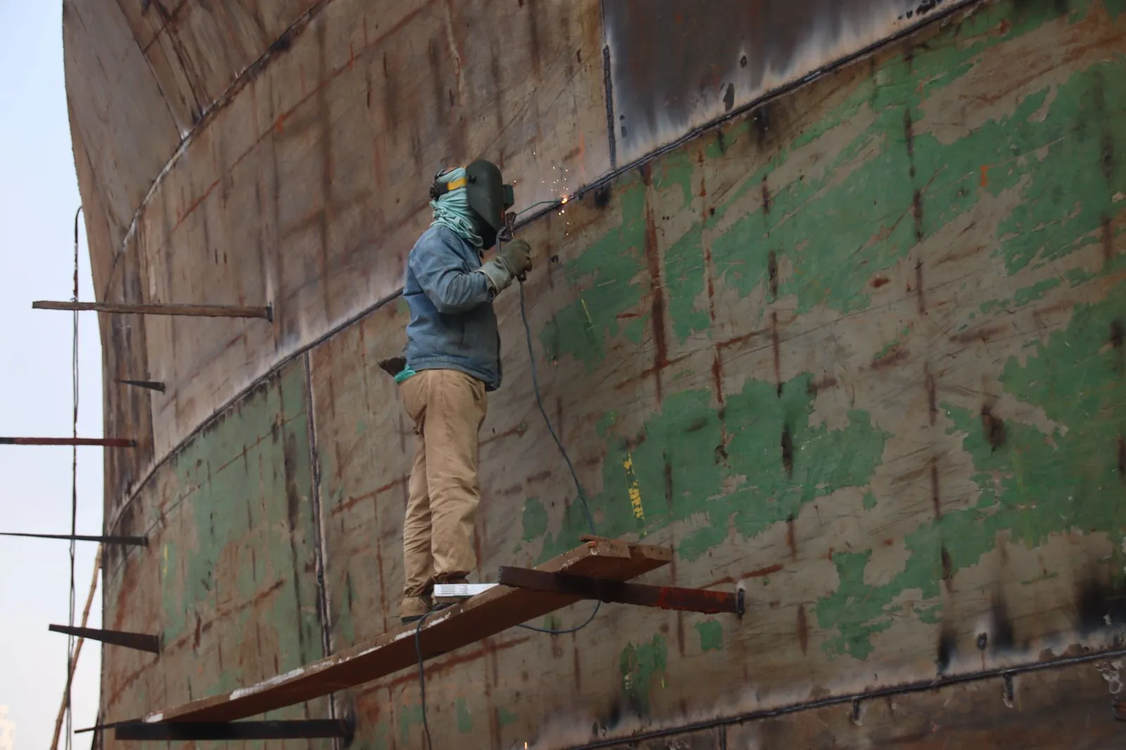 Welder on scaffolding running a weld bead on the curved shell of a large welded steel assembly.