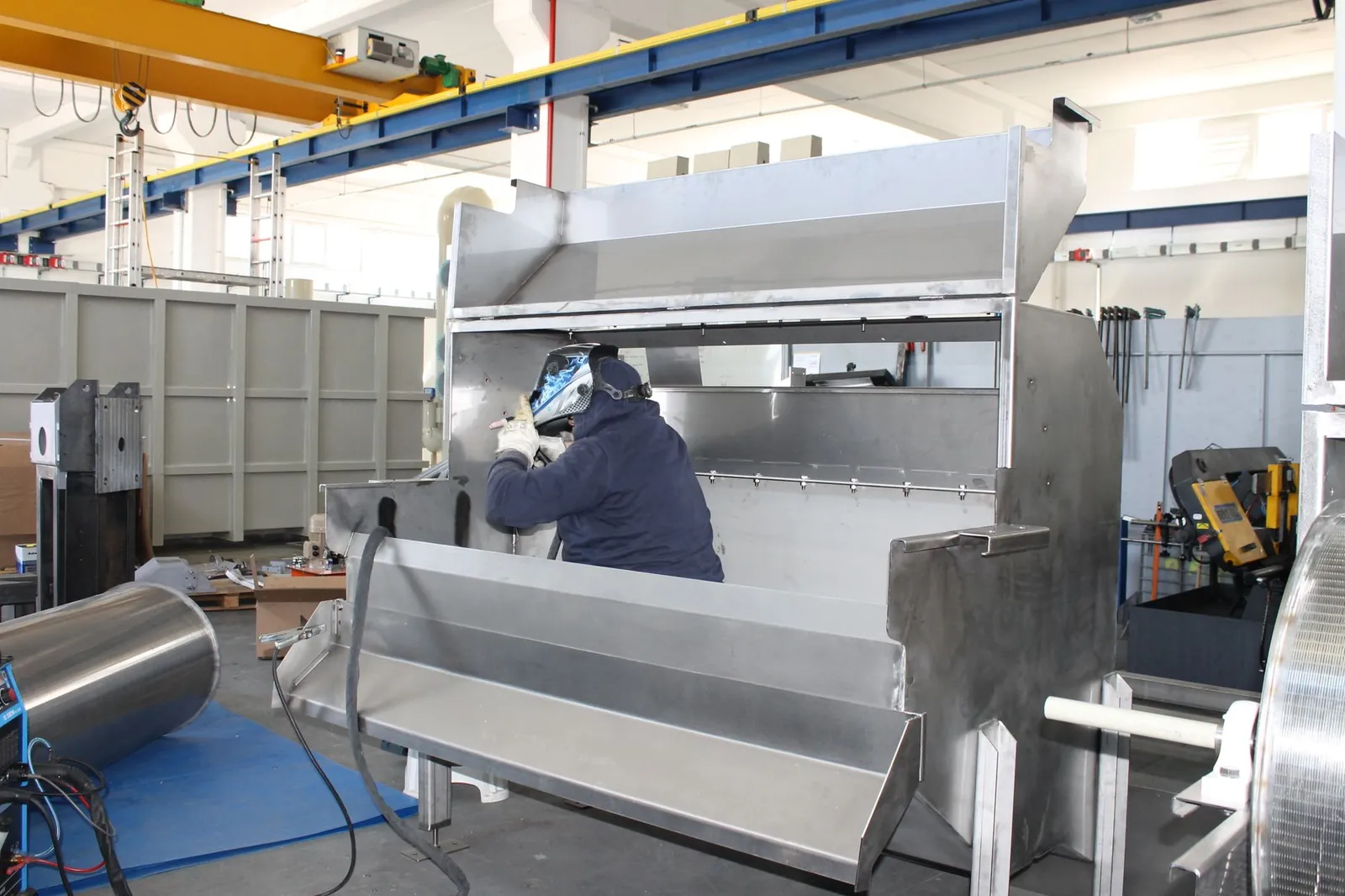 Welder assembling a large custom stainless steel weldment in a shop equipped with an overhead crane.