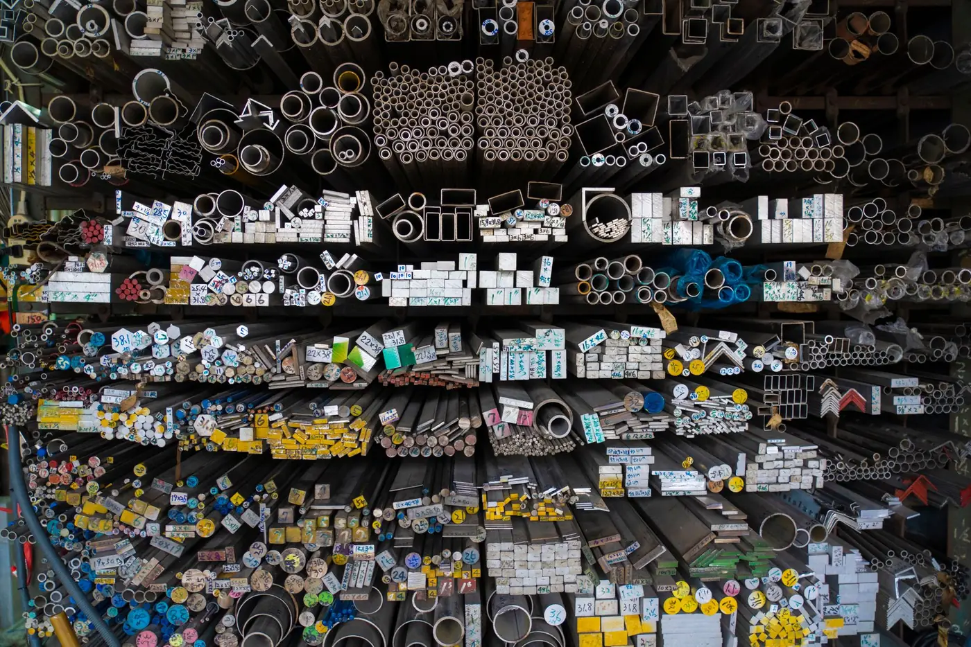 Rack of metal bars, tubes and profiles labeled with their dimensions, ready for incoming material inspection.